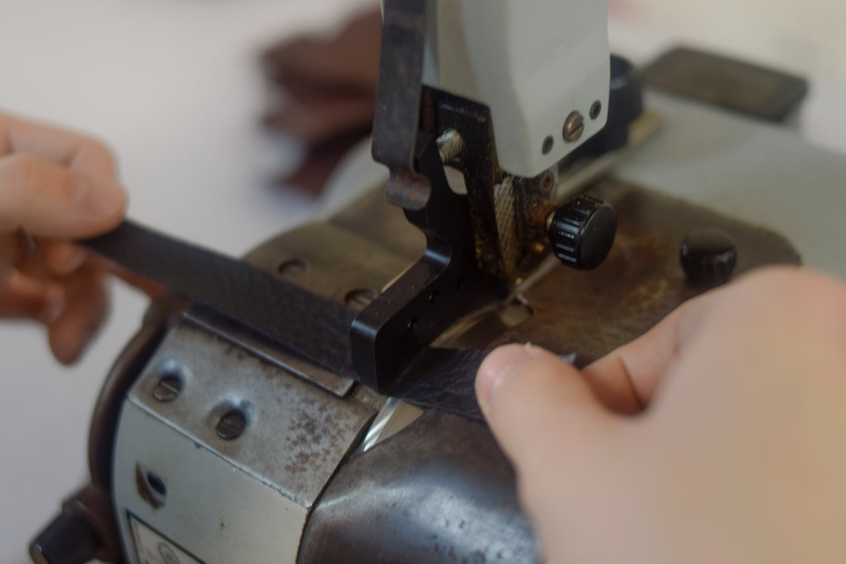 A close-up of a person's hands guiding a black leather strip through a vintage industrial sewing machine with a white and dark metal body.