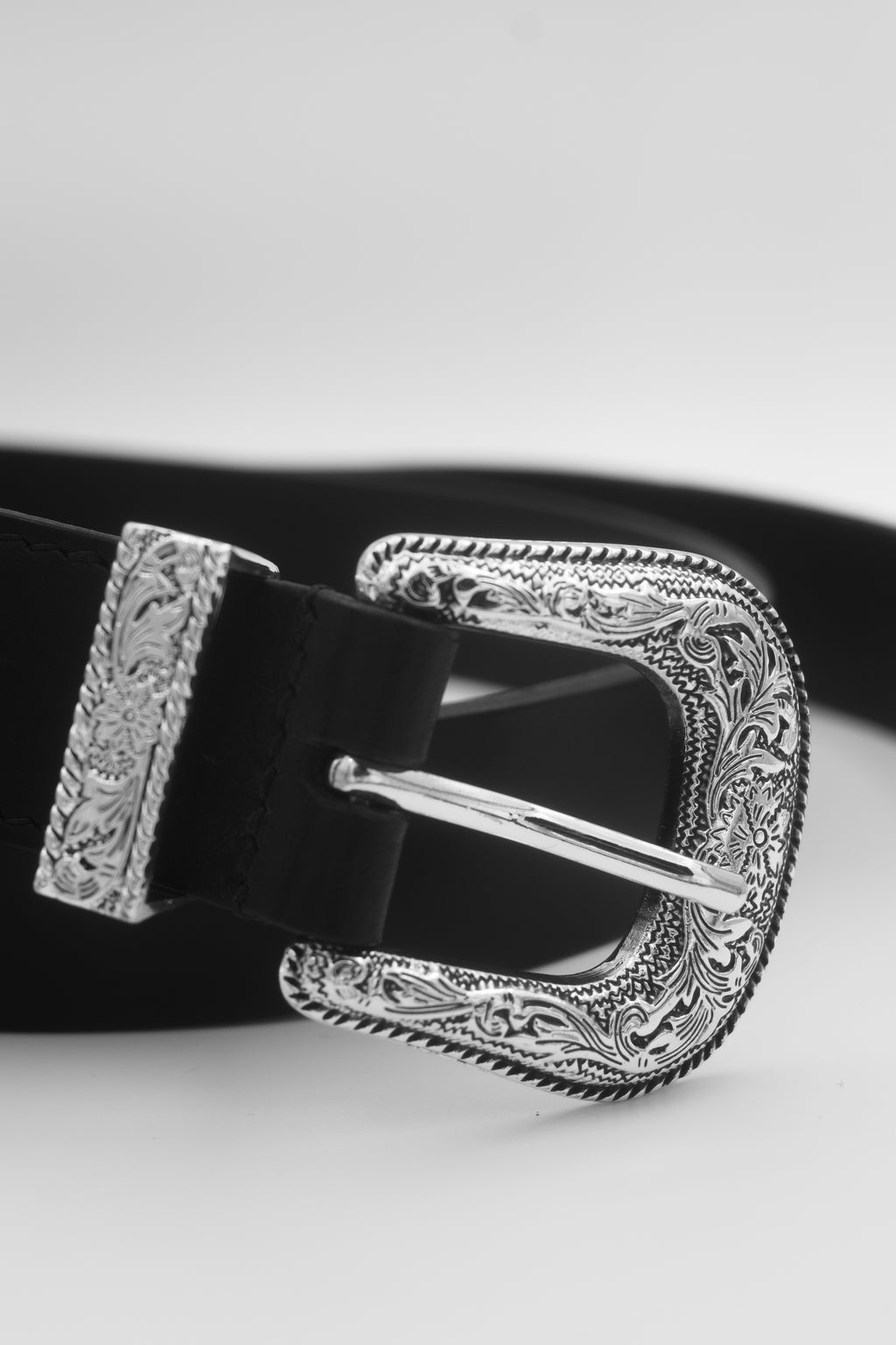A close-up of a black leather belt with an ornate, engraved silver-tone buckle and keeper, set against a clean light gray background.