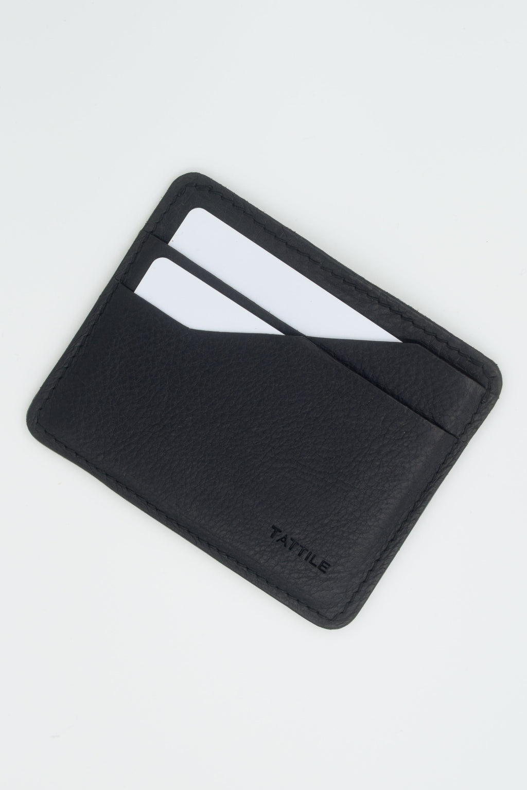 A black textured leather cardholder wallet with two card slots containing white cards, shown on a light gray background. The brand name 'TATILE' is debossed on the bottom right corner.